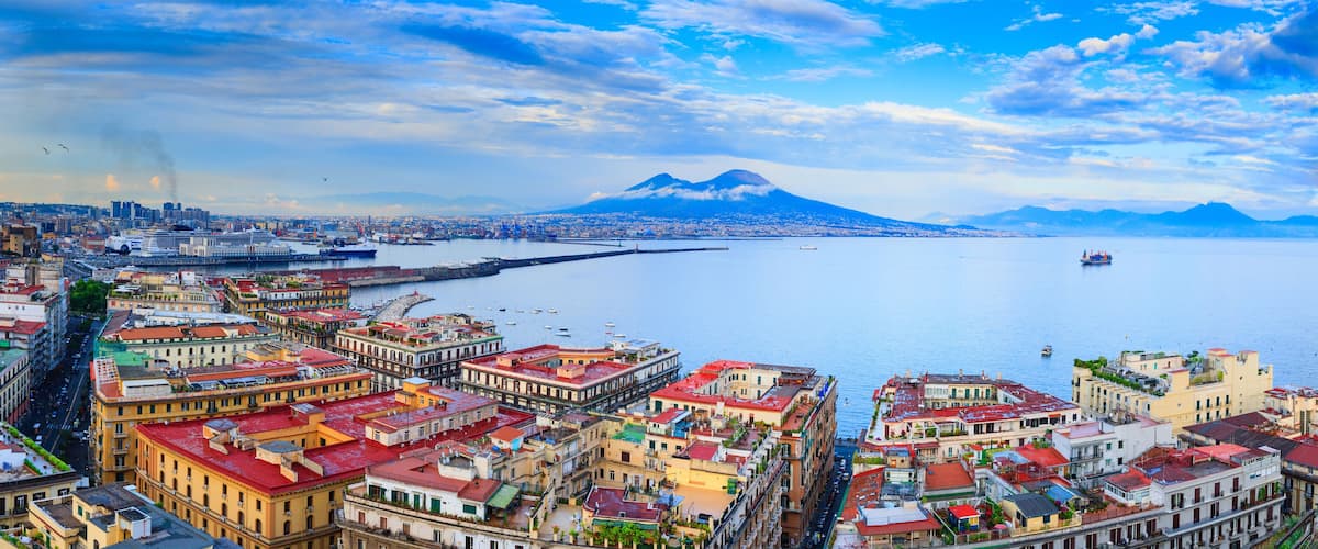 A coastal cityscape with historic buildings, a prominent mountain in the background, and a calm sea.