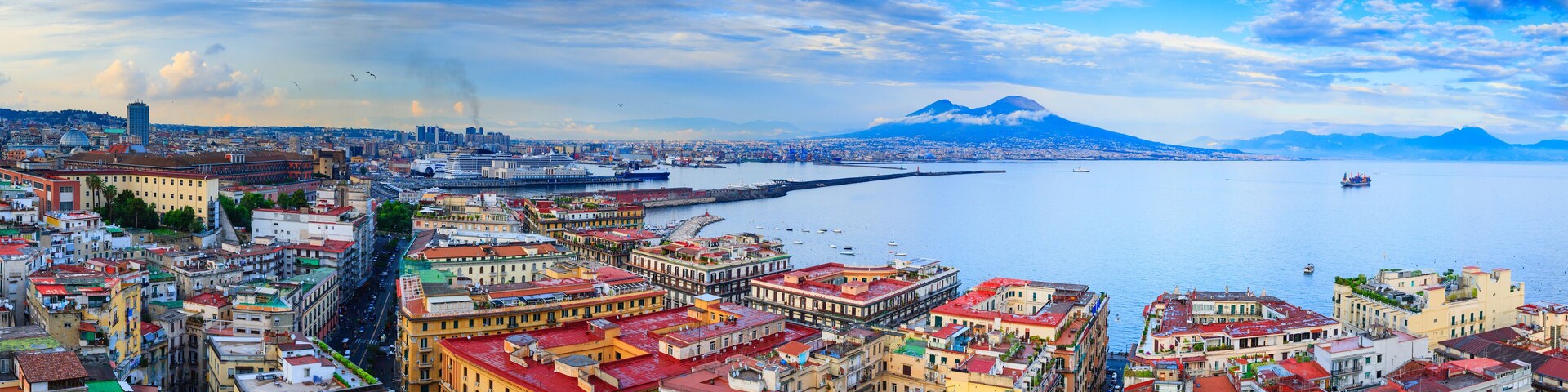 A coastal cityscape with historic buildings, a prominent mountain in the background, and a calm sea.