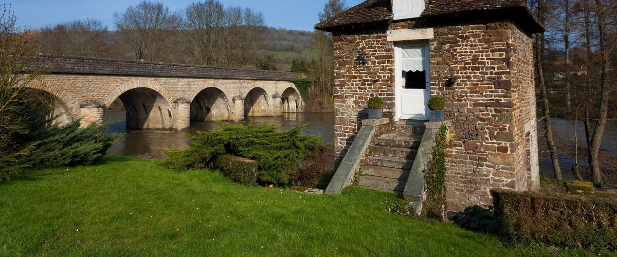 A stone bridge over a river, a small stone building with a sloped roof, and a grassy area with bushes.