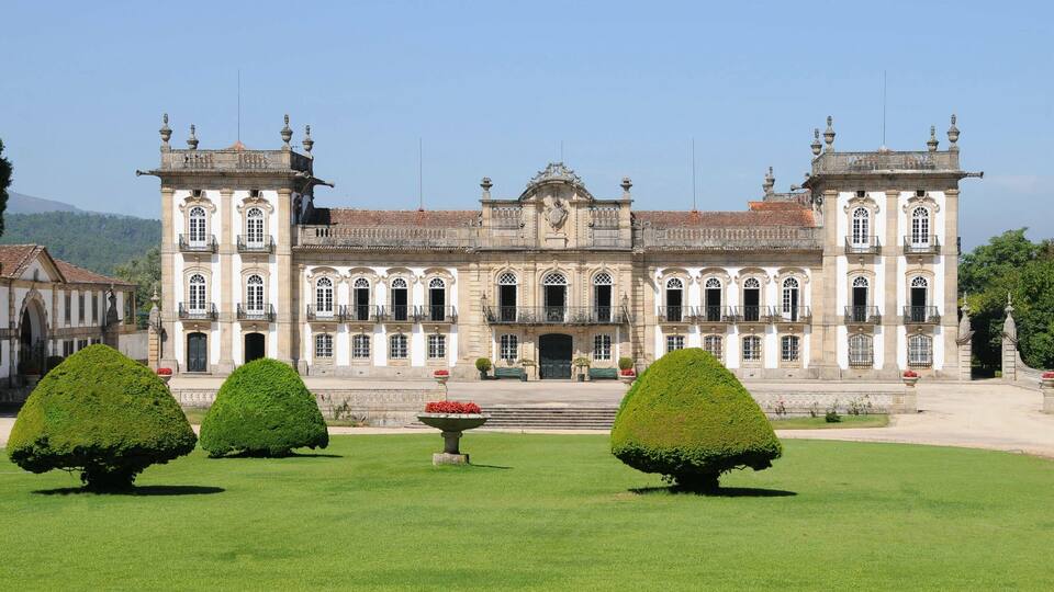 A large, historic building with symmetrical architecture, a central entrance, and a well-maintained garden with trimmed hedges and flower beds.