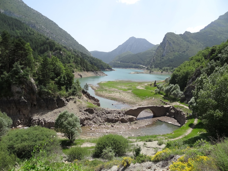 A river flowing through a valley with a stone bridge, surrounded by lush green hills and a mountain in the background.