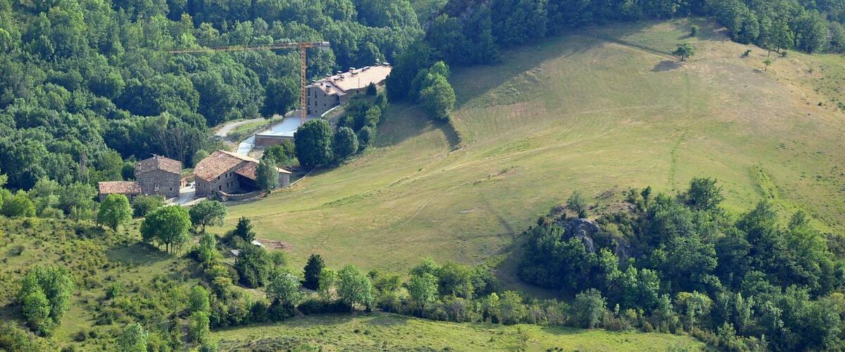 A rural landscape with rolling hills, a small cluster of buildings, and dense forested areas.