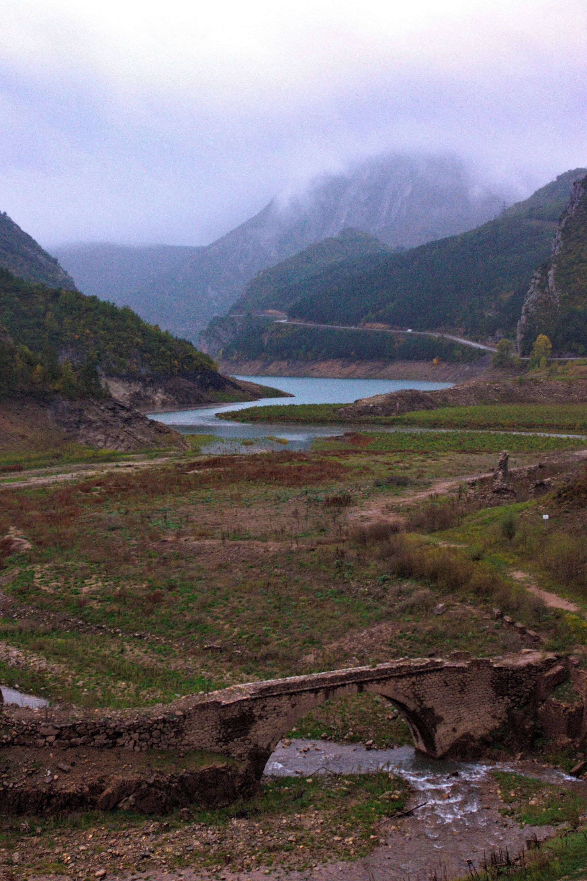 A stone bridge over a river, surrounded by mountains and a lake.