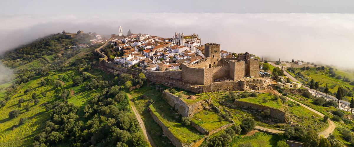 A historic town with a prominent castle on a hill, surrounded by lush greenery and a sea of clouds.