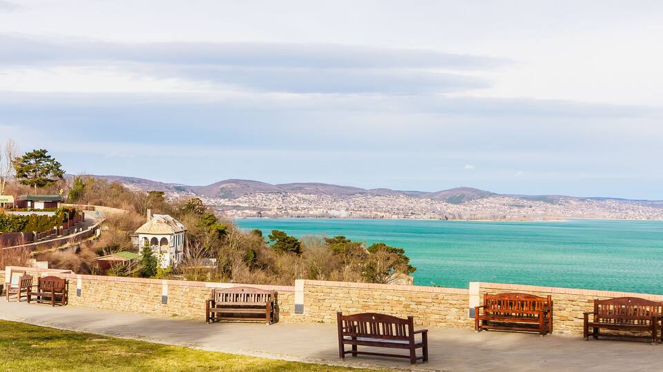A coastal cityscape with a clear view of the sea, stone benches, and a distant hill.