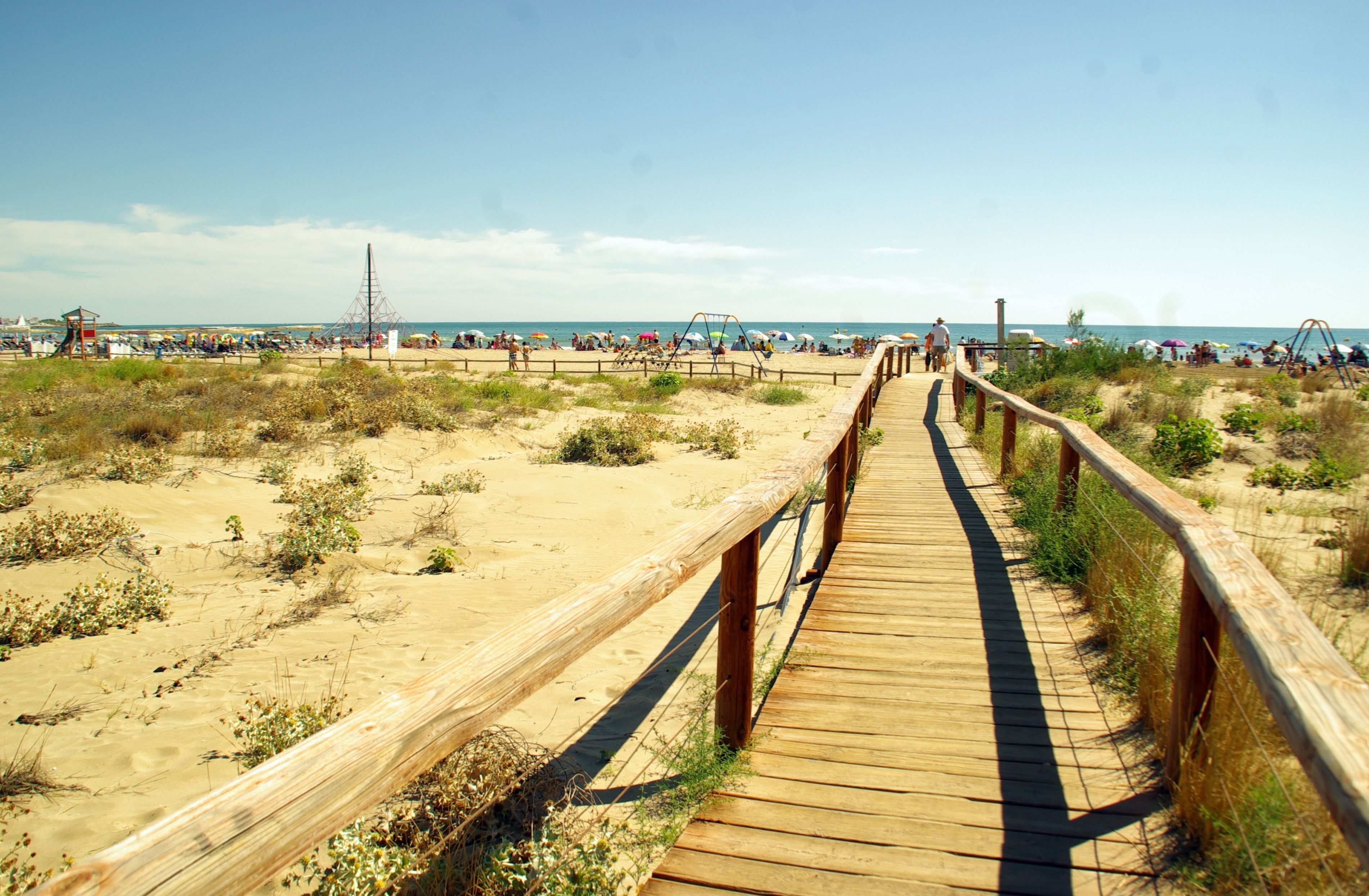 Una pasarela de madera que lleva a una playa arenosa, con gente y sombrillas.