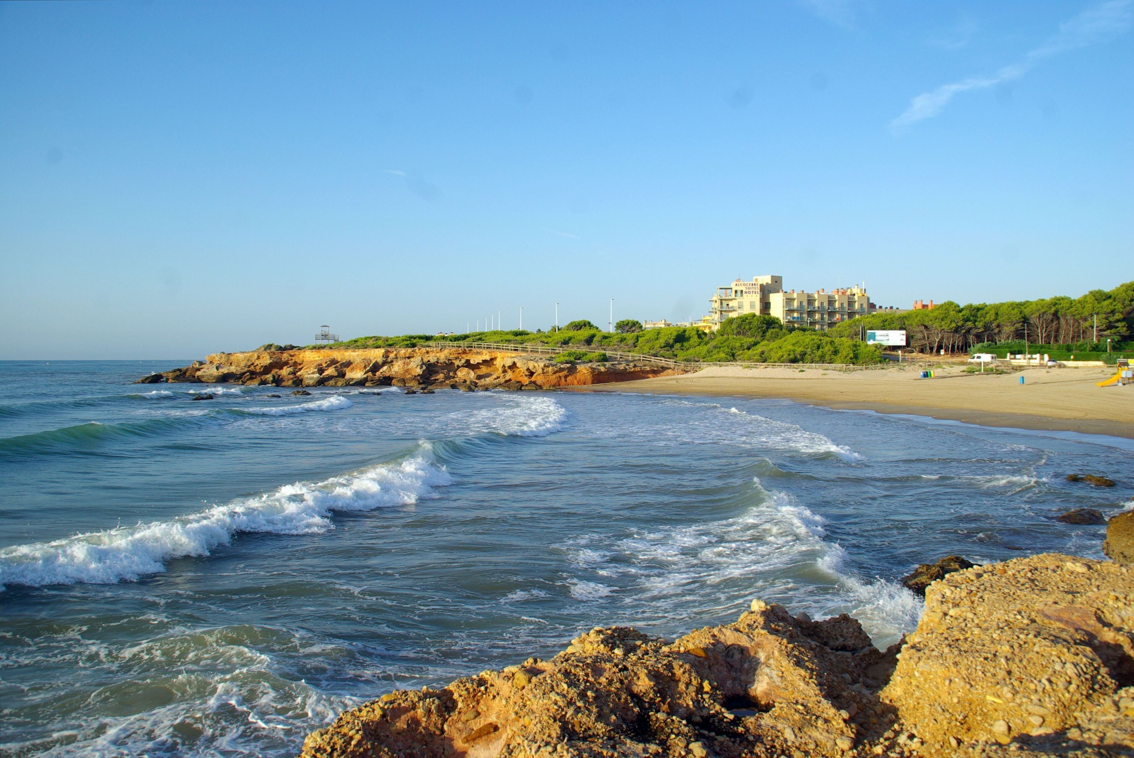 Una playa costera con olas, costa rocosa y un edificio al fondo.