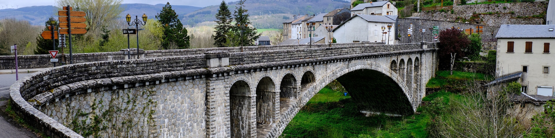 Uma ponte de pedra com várias arcos sobre um rio, rodeada por vegetação e edifícios.