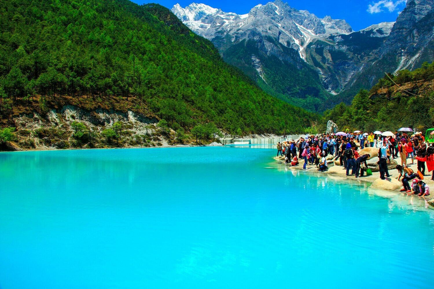 A turquoise lake surrounded by green mountains and a crowd of people.
