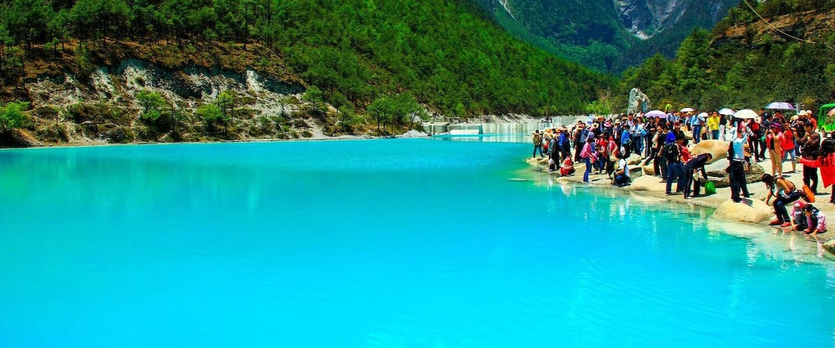 A turquoise lake surrounded by green mountains and a crowd of people.