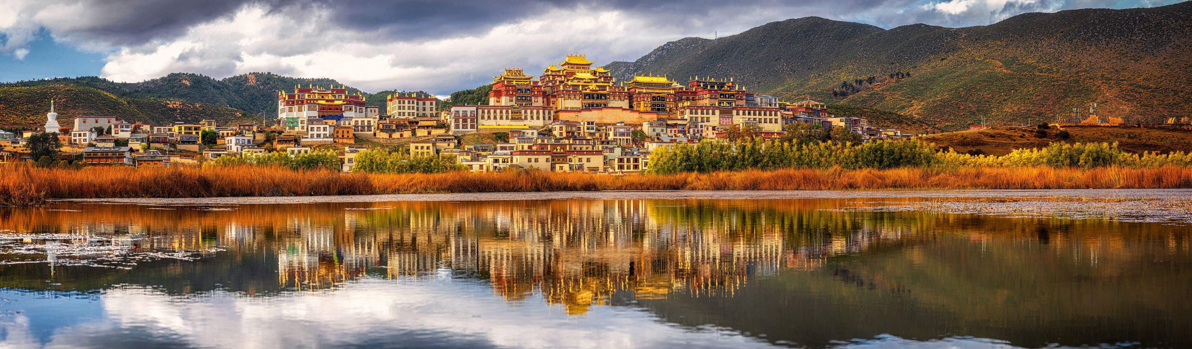 A mountainous region with a cluster of colorful buildings, a reflective lake, and a prominent white stupa.