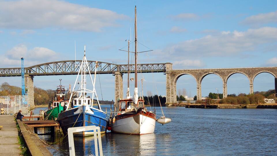A river with boats docked, a bridge, and stone arches in the background.