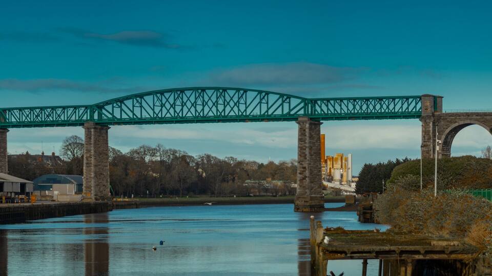 A green steel bridge over a river with stone arches, surrounded by industrial buildings and a clear sky.
