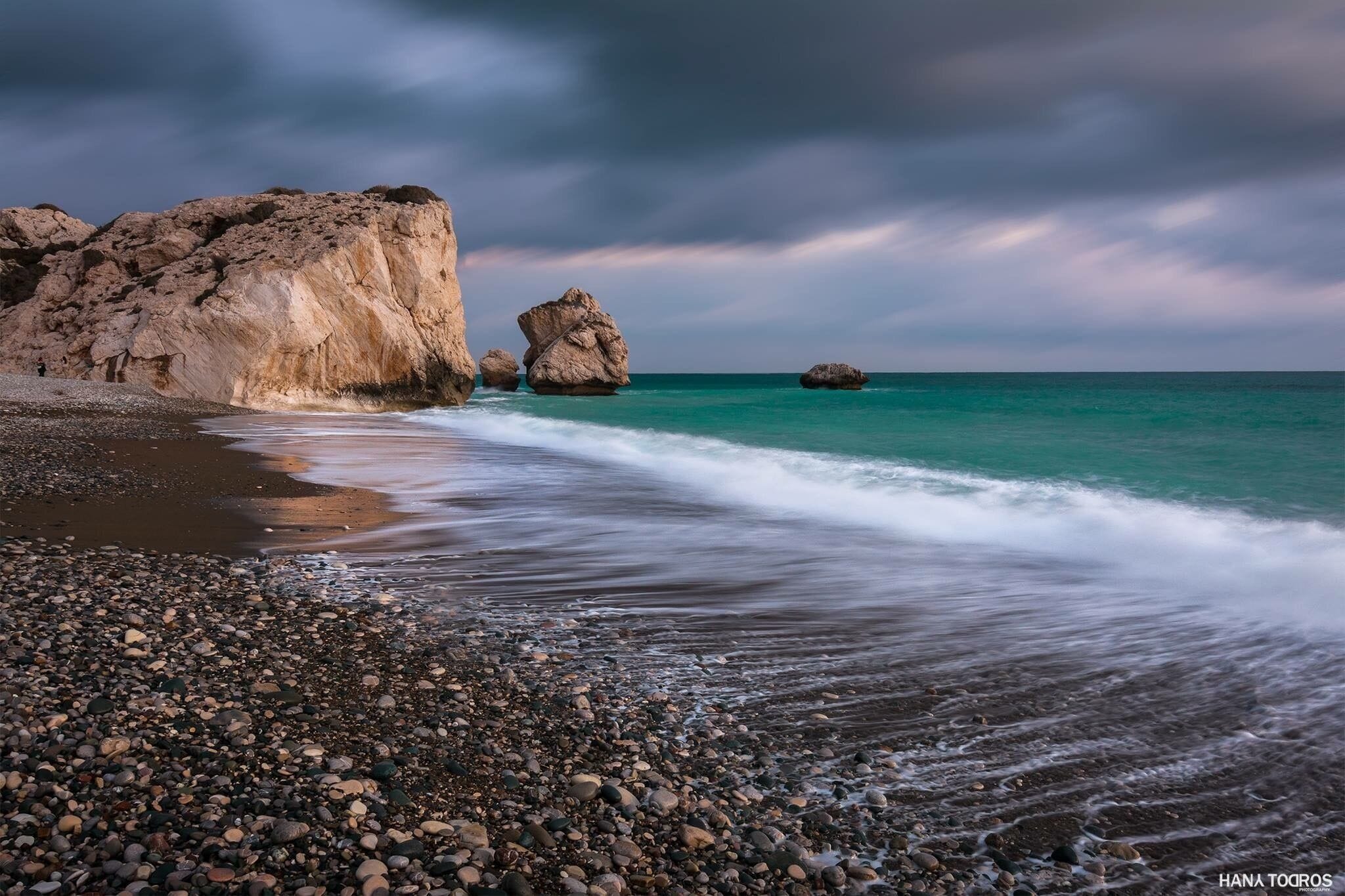 A rocky beach with waves crashing onto the shore and a large rock formation in the background.