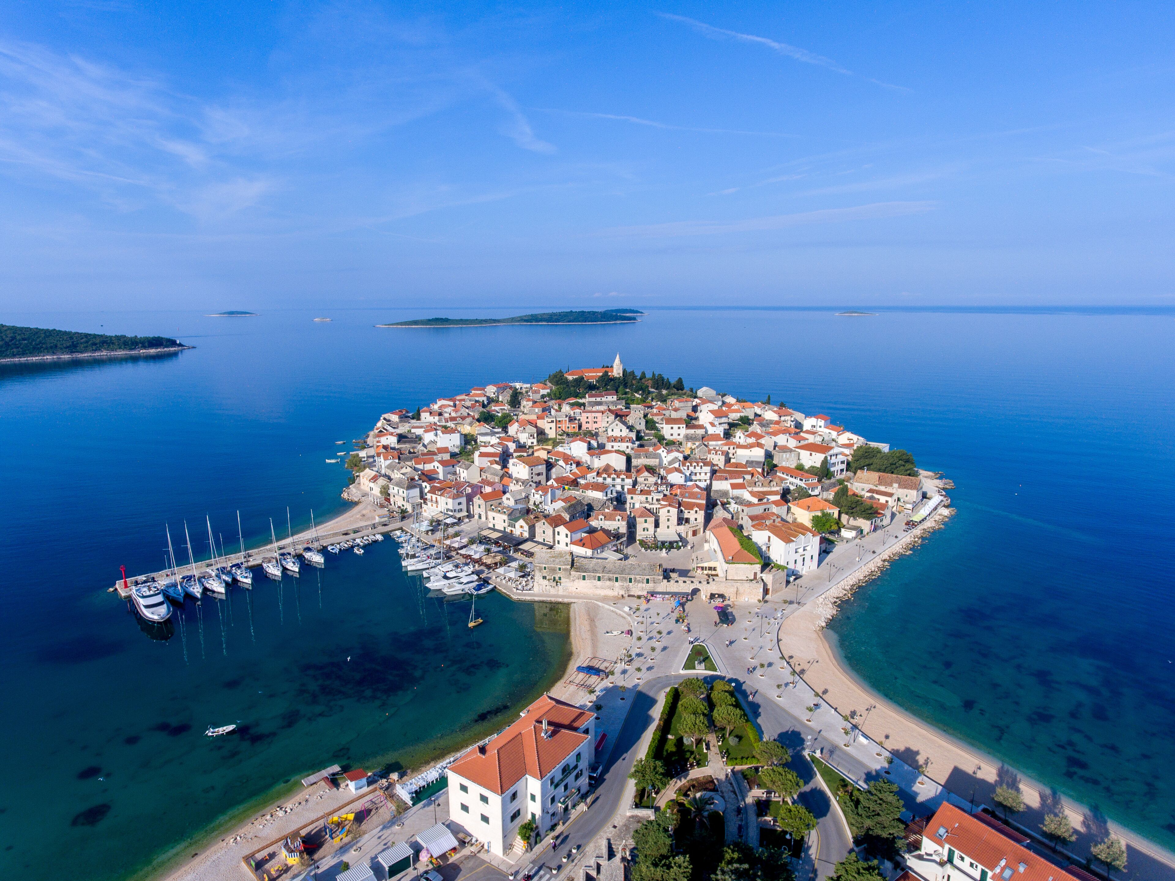An aerial view of a coastal town with a central island, a marina, and clear blue waters.
