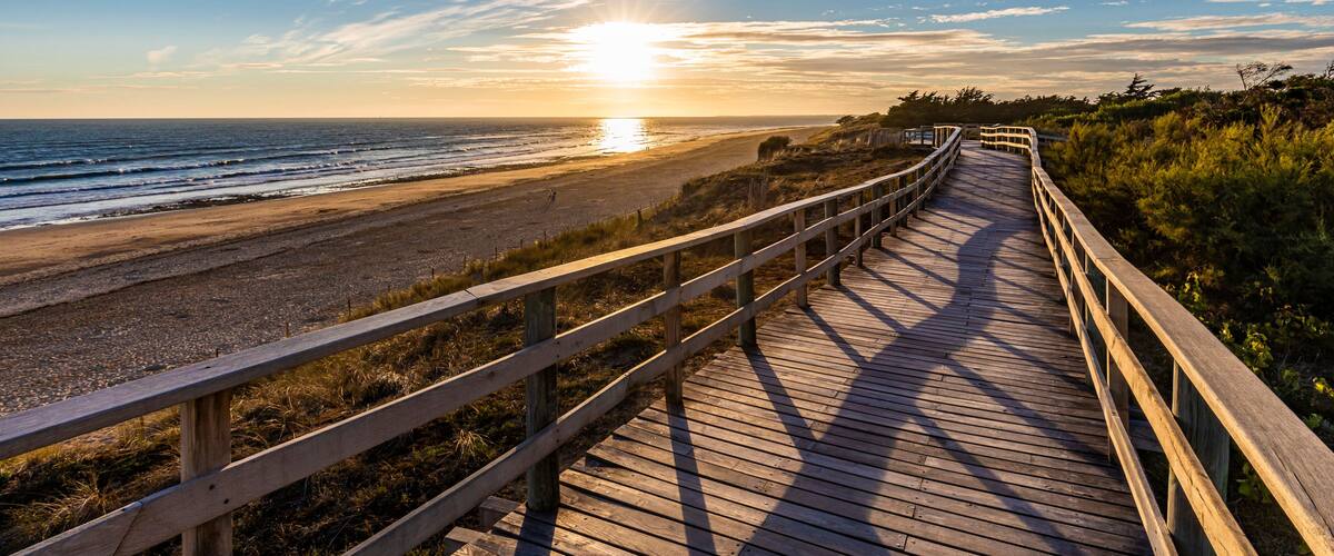 A wooden boardwalk along a beach with the ocean and sunset in the background.