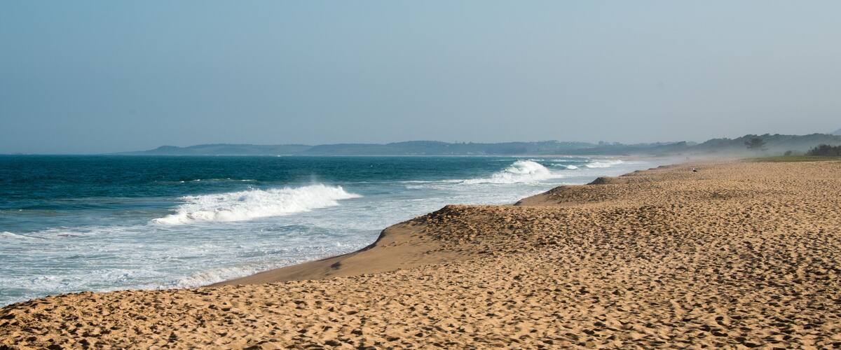 A sandy beach with waves crashing onto the shore, a clear sky, and distant hills.