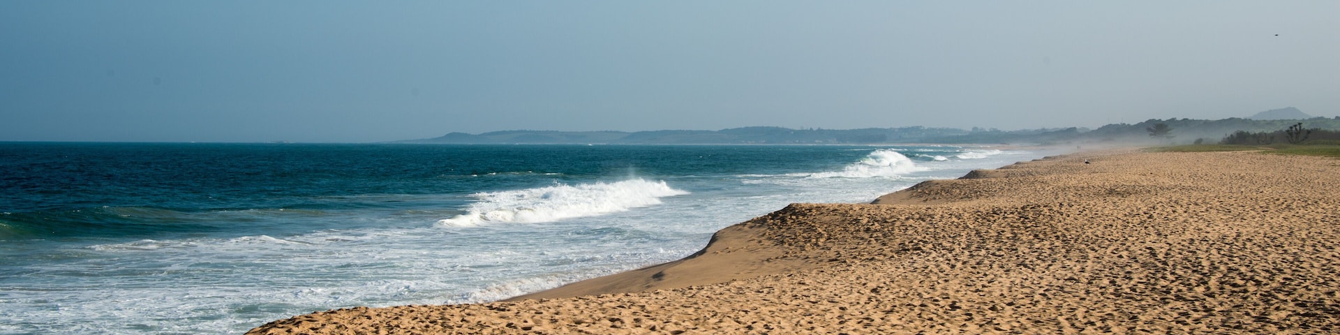 A sandy beach with waves crashing onto the shore, a clear sky, and distant hills.