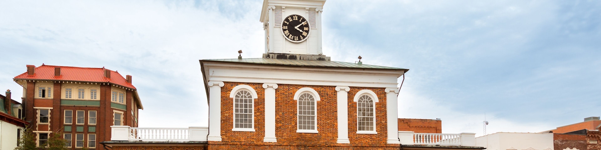 A historic clock tower with a white clock face, surrounded by brick buildings and a roundabout.