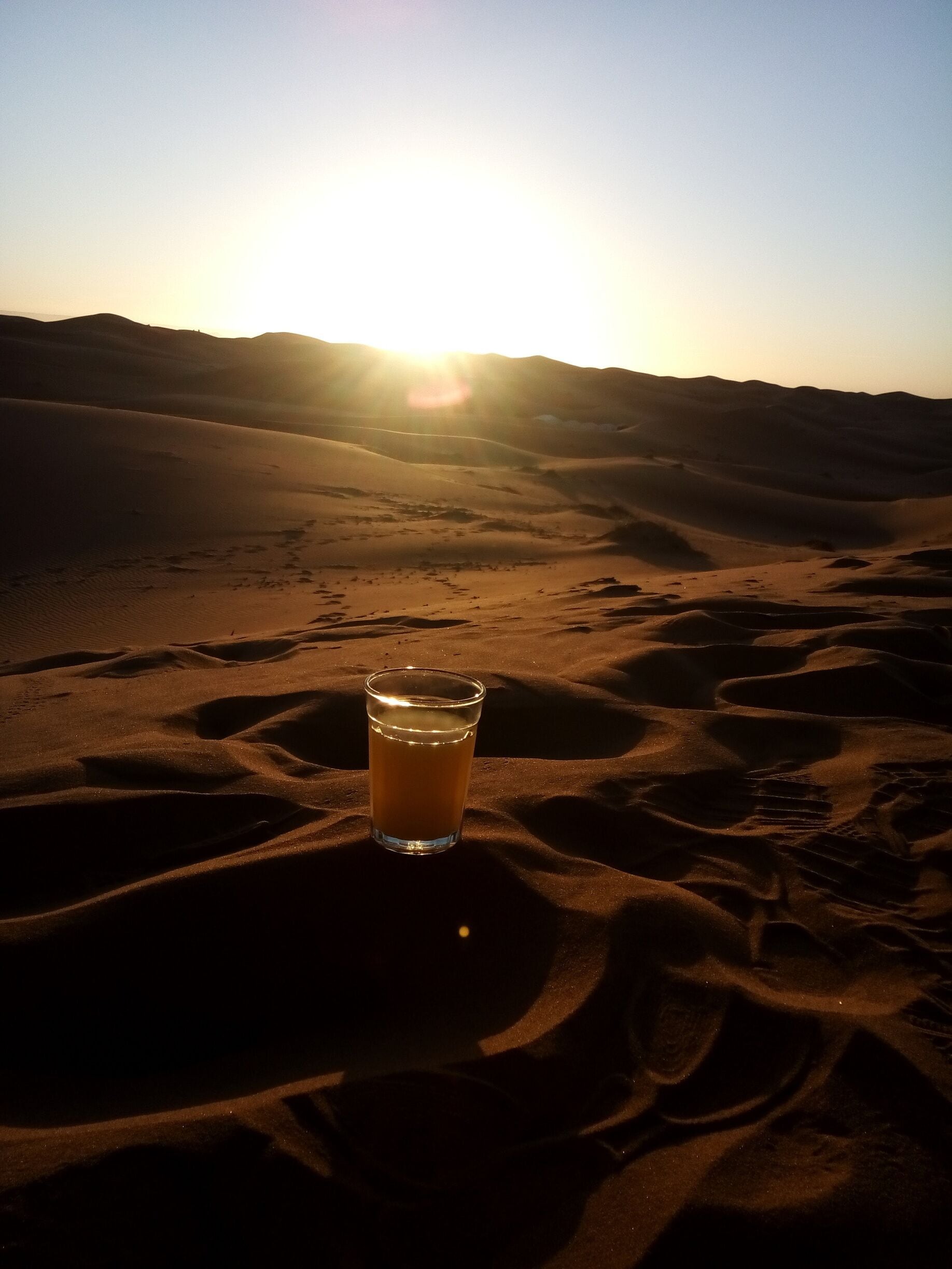 A glass of drink on sand dunes with the sun setting in the background.