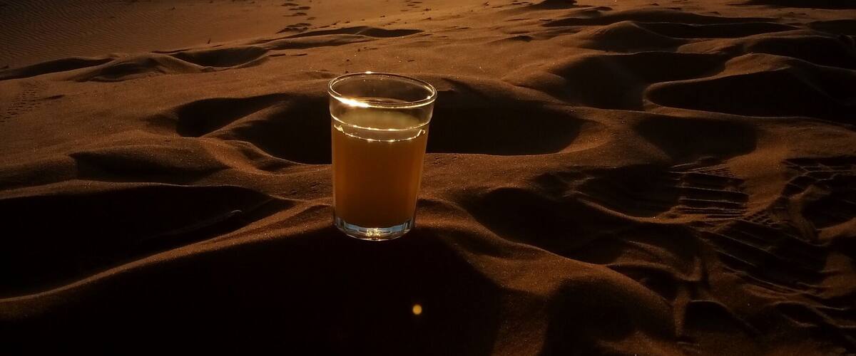A glass of drink on sand dunes with the sun setting in the background.