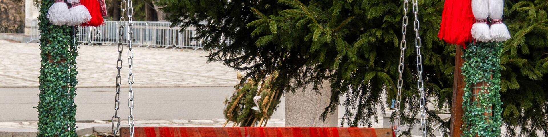 An outdoor wooden swing elaborately decorated with traditional red and white Martenitsa figures and green foliage, featuring a 'Chestitia Baba Marta' sign in Razlog.