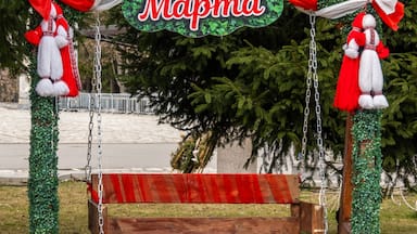 An outdoor wooden swing elaborately decorated with traditional red and white Martenitsa figures and green foliage, featuring a 'Chestitia Baba Marta' sign in Razlog.