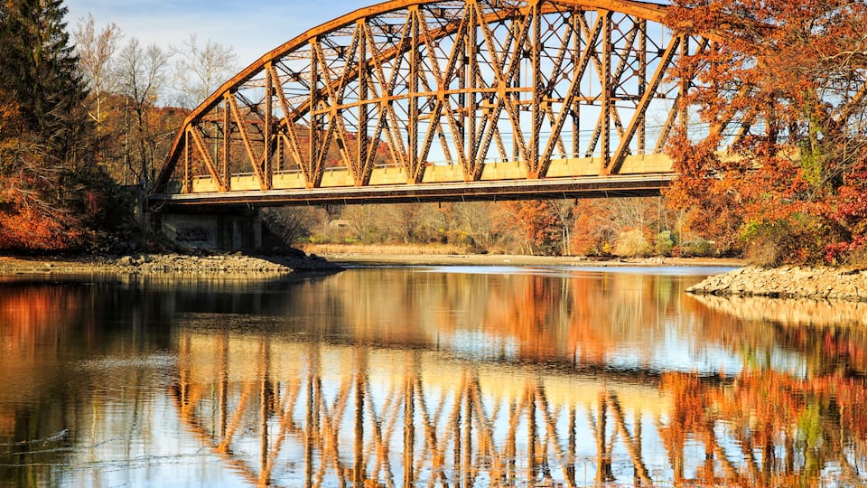 A steel bridge over a calm river with autumn foliage.