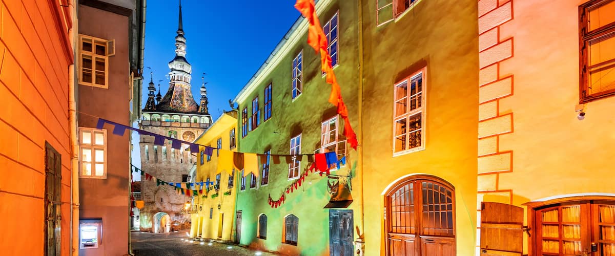A cobblestone street lined with colorful buildings, a prominent tower, and flags.