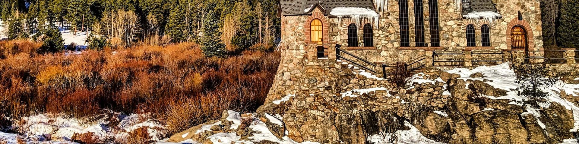 A stone church with a cross on top, surrounded by snow-covered mountains and trees.