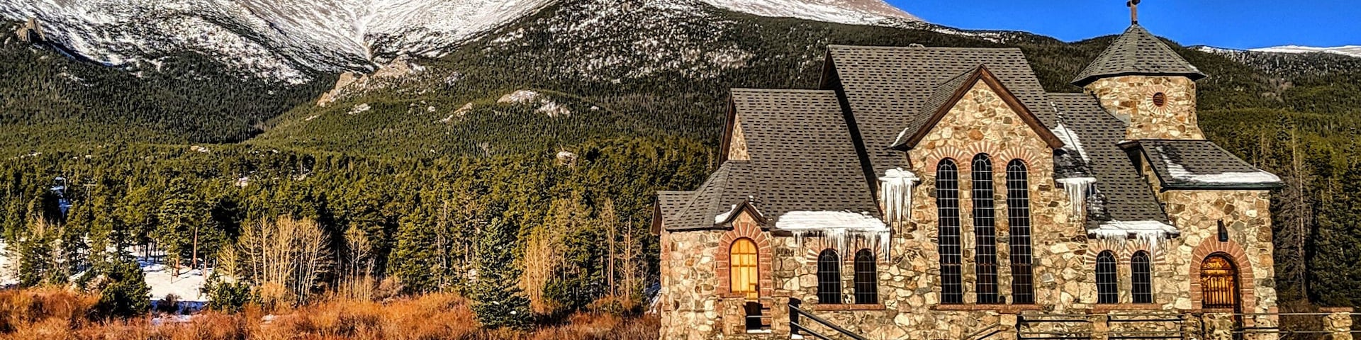 A stone church with a cross on top, surrounded by snow-covered mountains and trees.