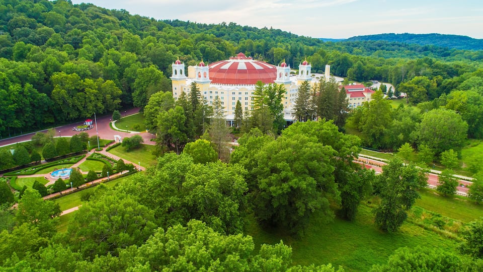 A large, historic building with a red roof, surrounded by lush green trees and hills.