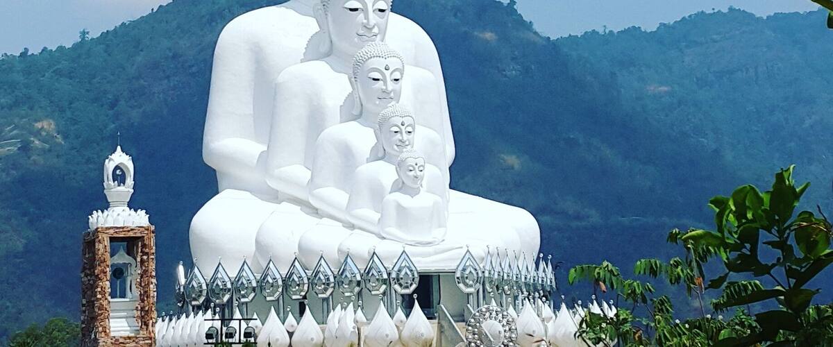 A large white Buddha statue with a golden spire, surrounded by smaller statues and a white fence with pointed tops, set against a mountainous backdrop.