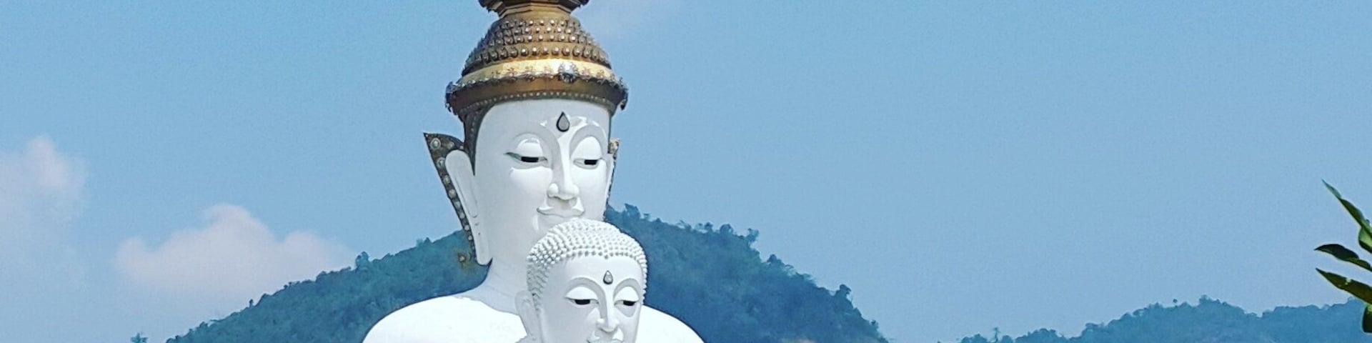 A large white Buddha statue with a golden spire, surrounded by smaller statues and a white fence with pointed tops, set against a mountainous backdrop.