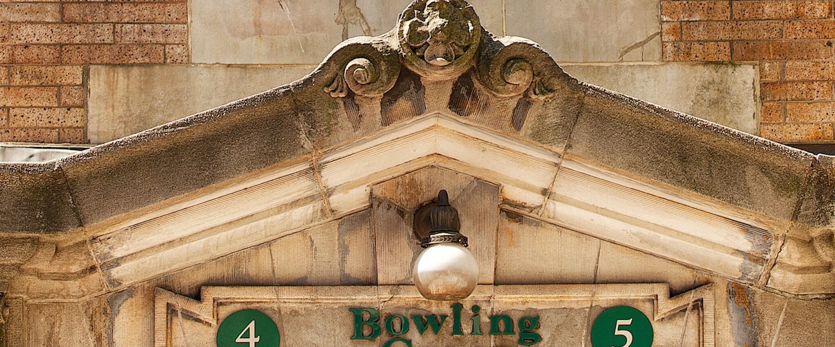 A clock with Roman numerals on a stone building, surrounded by decorative elements and a sign indicating "Bowling Green."
