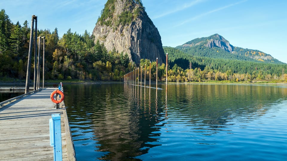 A wooden pier extending into calm waters, a rocky mountain in the background, and a forested hillside.