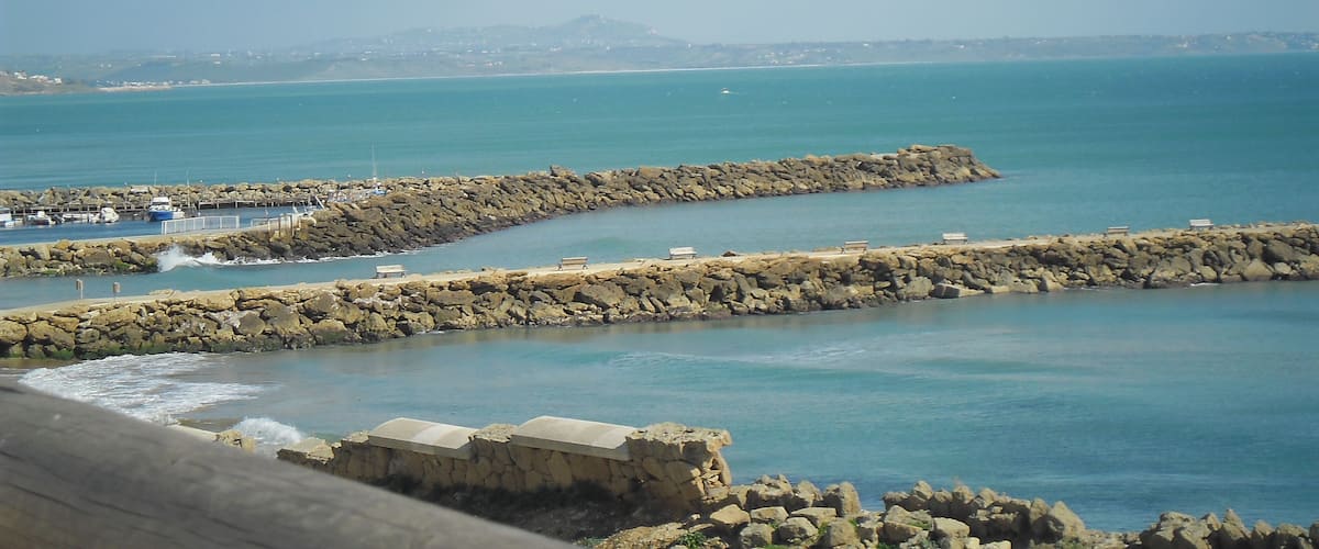 A coastal view with a rocky breakwater, calm sea, and distant hills.