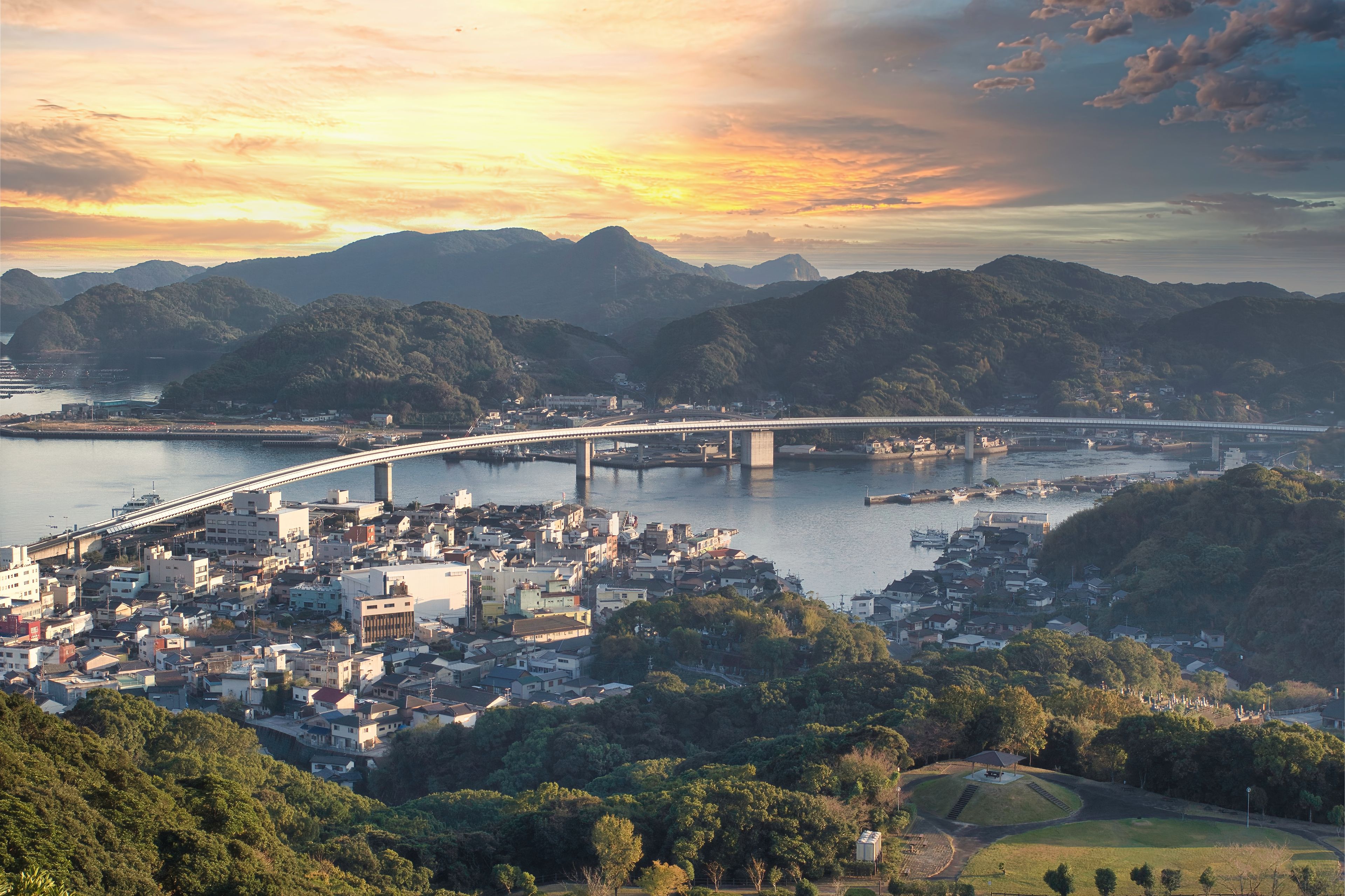 A coastal cityscape with a prominent bridge spanning a river, surrounded by lush hills and a sunset sky.