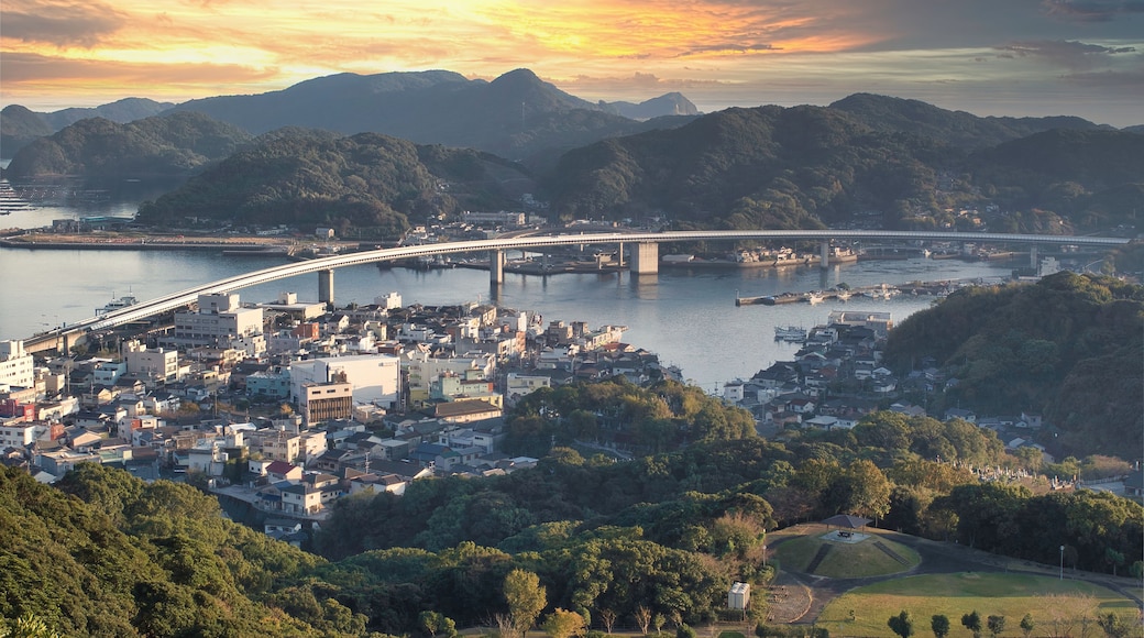 A coastal cityscape with a prominent bridge spanning a river, surrounded by lush hills and a sunset sky.