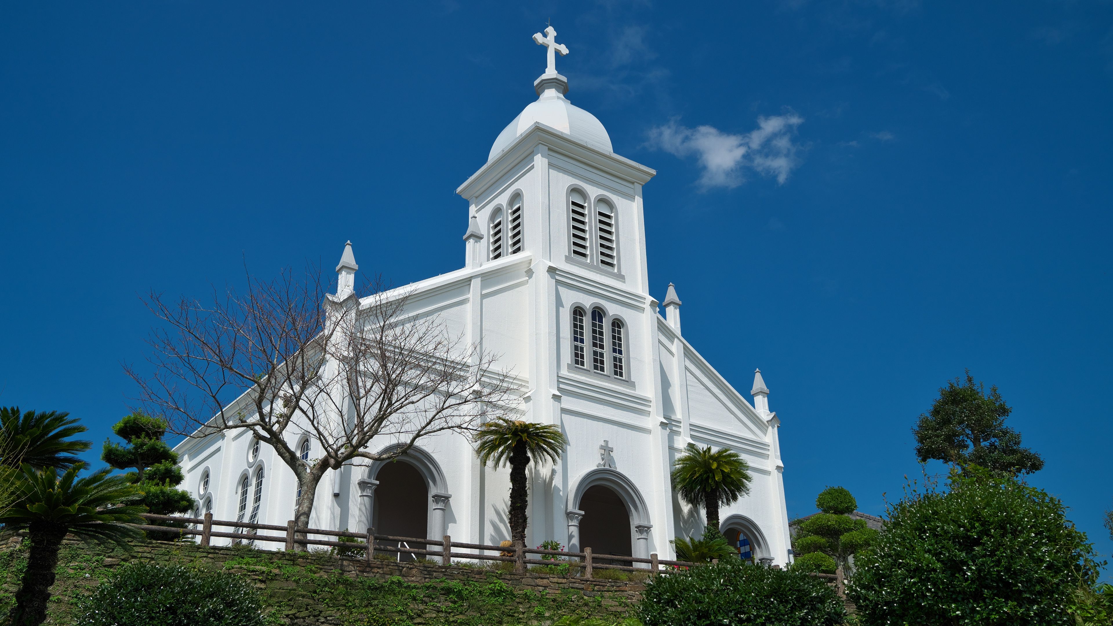 A white church with a cross on top, surrounded by greenery and a clear blue sky.