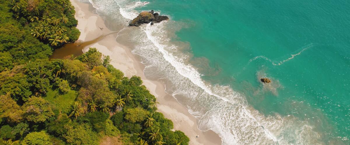 An aerial view of a beach with clear turquoise waters, sandy shore, and dense tropical vegetation.