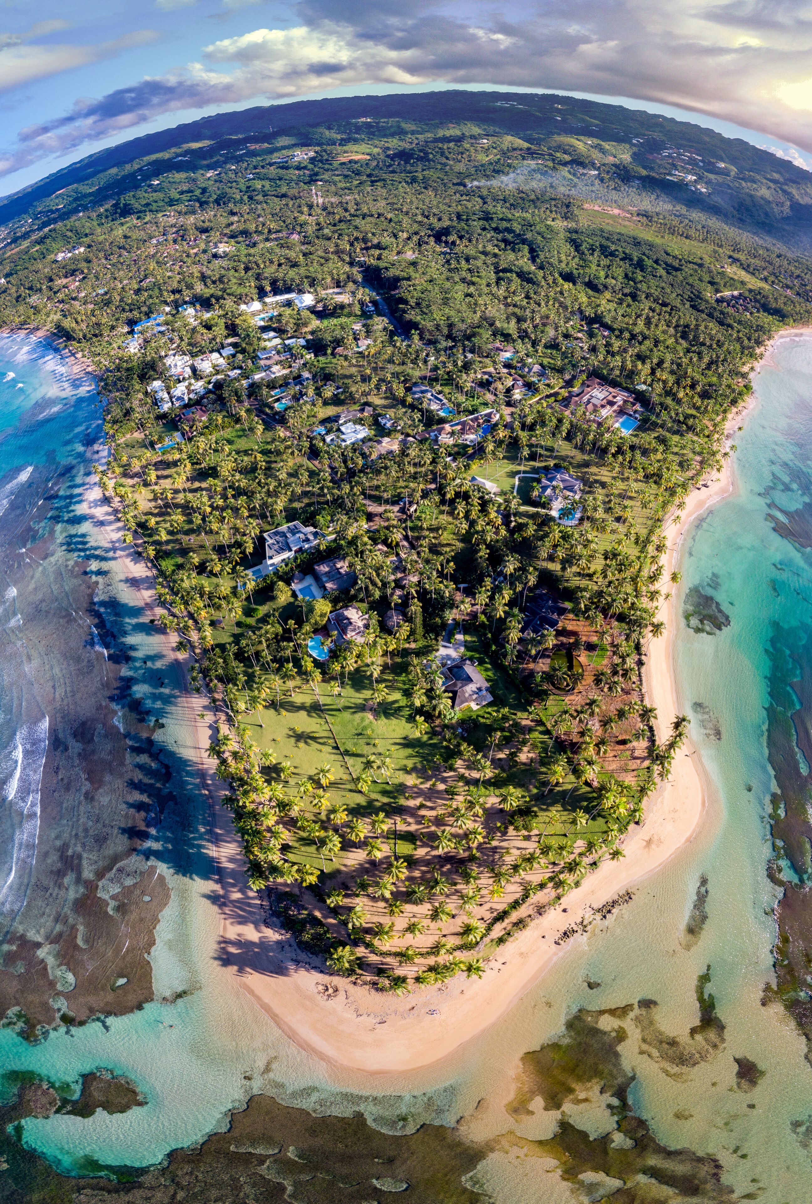Vista aérea de una zona costera con playa, aguas turquesas cristalinas y un denso conjunto de casas rodeadas de vegetación.
