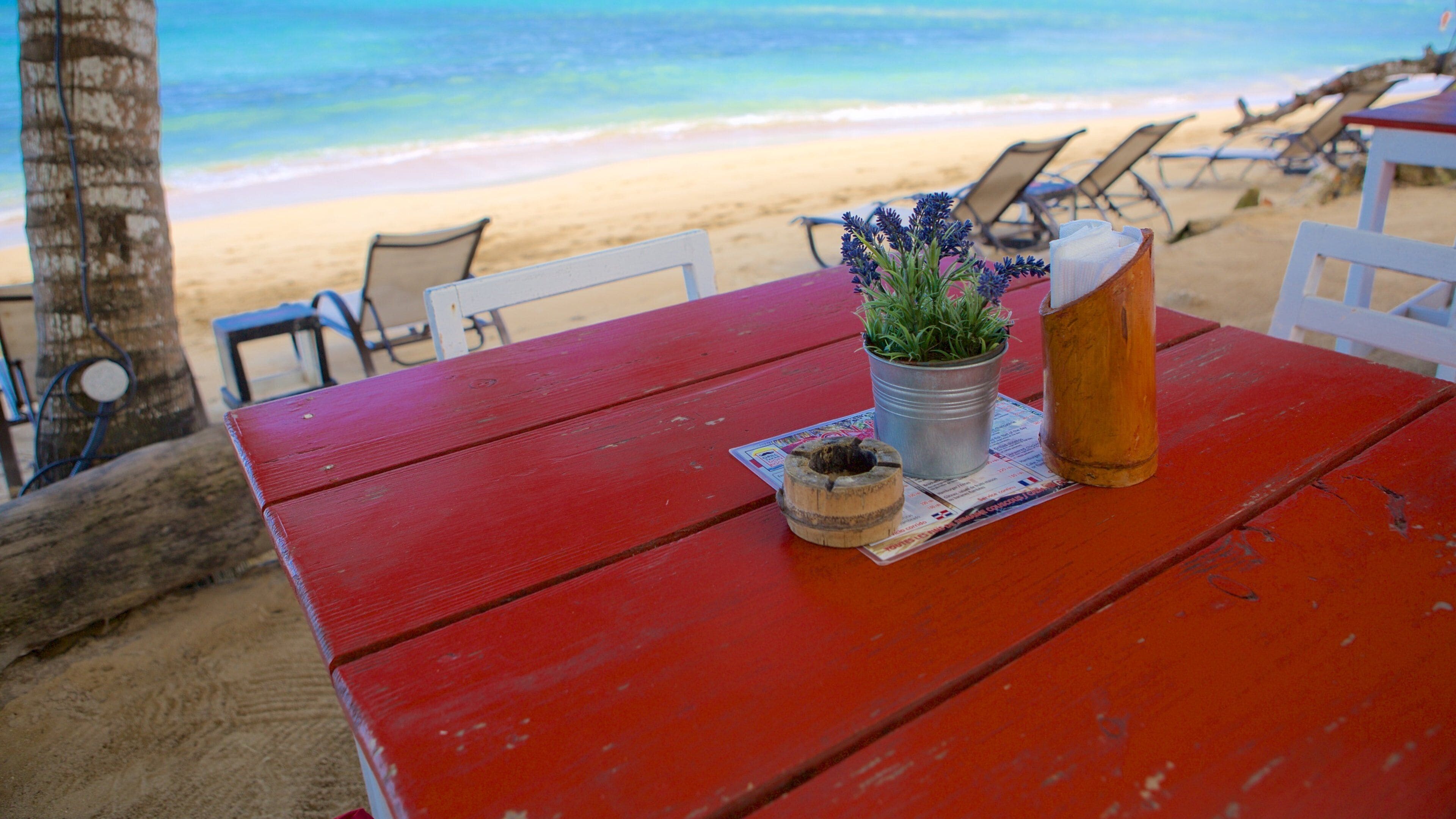 A red wooden table with a potted plant, a candle, and a menu on a beach with chairs and a palm tree.