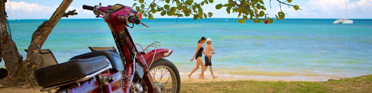 Une moto est garée sur une plage de sable, offrant une vue dégagée sur l’océan et on aperçoit un voilier au loin.