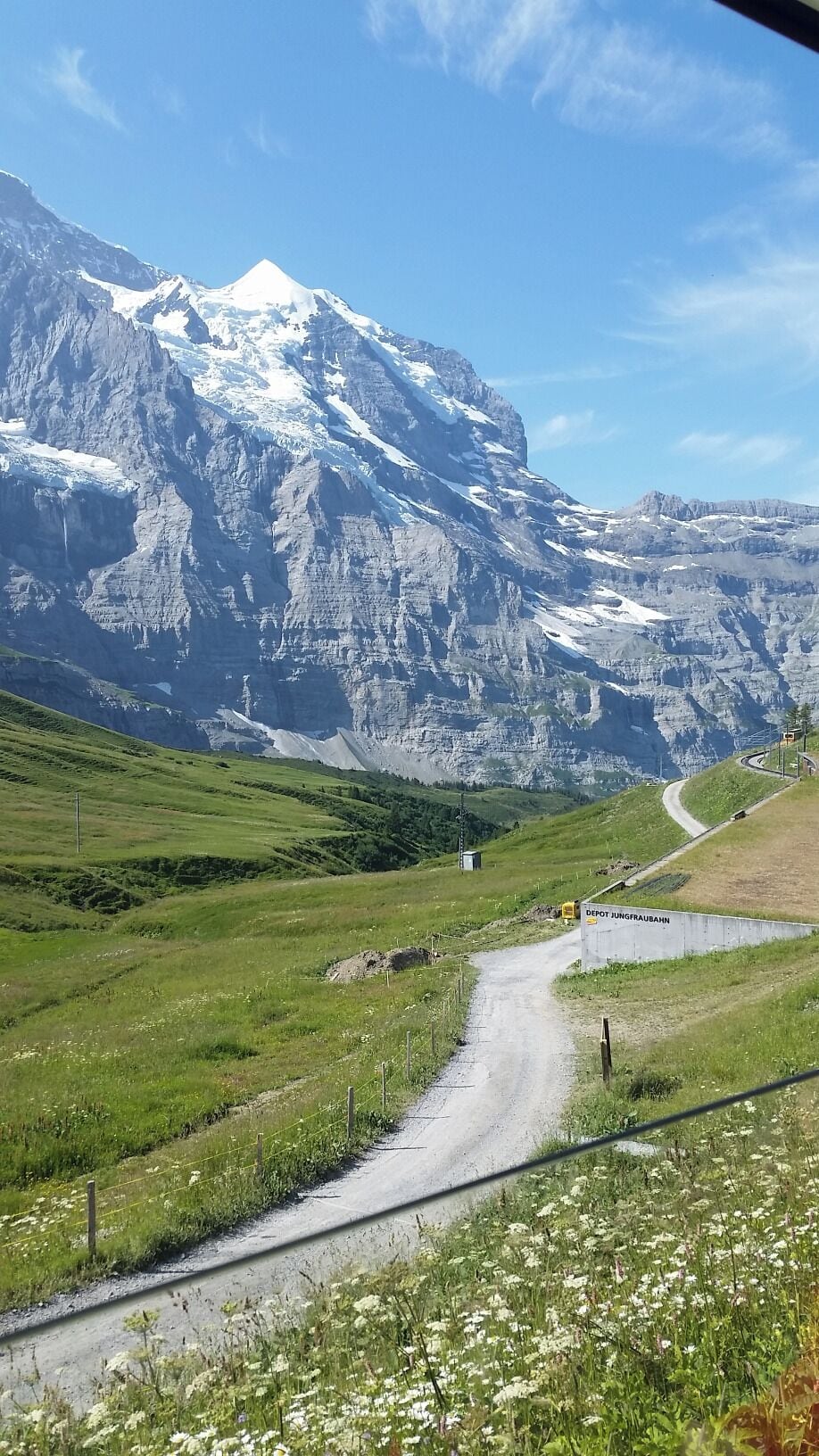 Eine Bergstraße mit einem Schild für Depot Jungfrauhahn, umgeben von schneebedeckten Gipfeln und grünen Feldern.