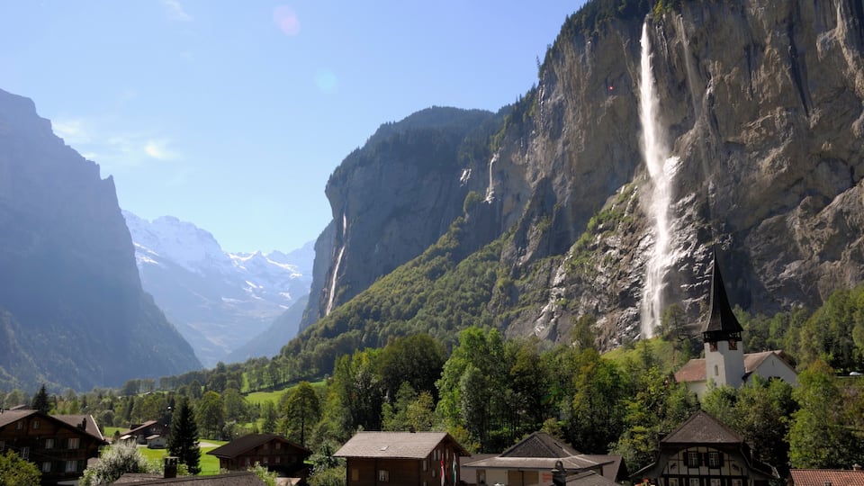A village with traditional houses, a church, and a waterfall in a mountainous valley.