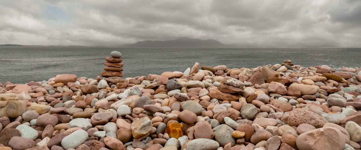 A rocky shoreline with a stack of stones in the foreground and a cloudy sky above.