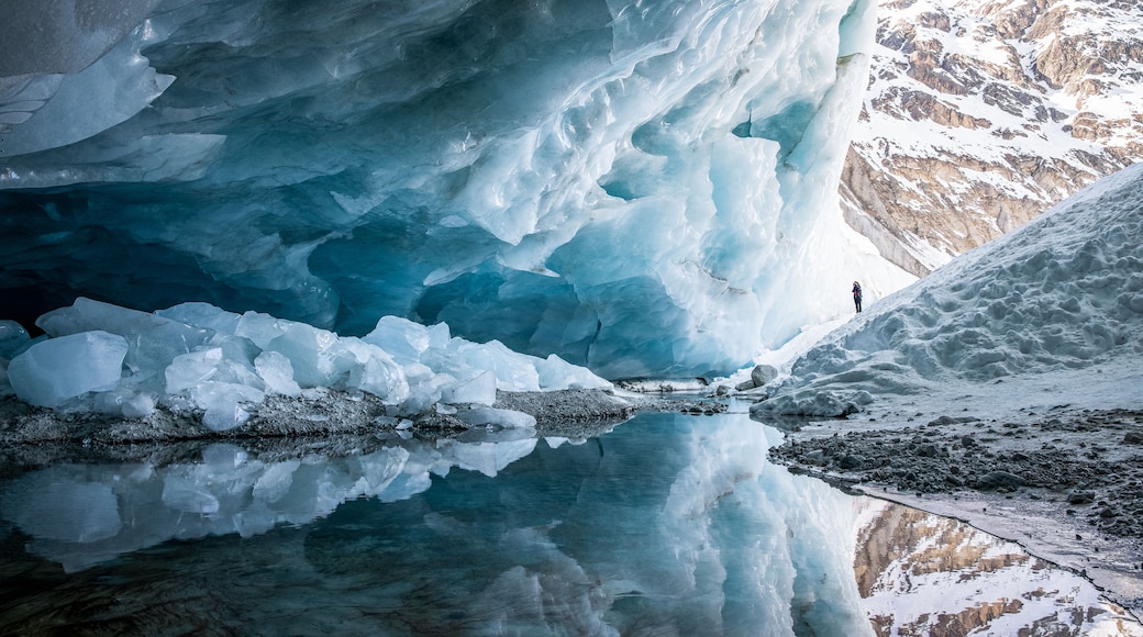 A glacier with a person standing at the edge, reflecting in a calm water body.