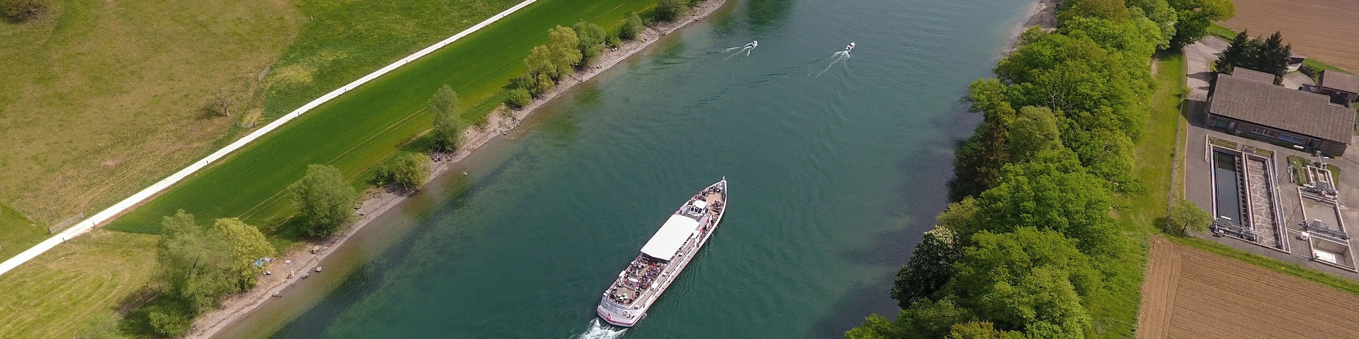 Um rio com um barco, rodeado por campos verdes e árvores.
