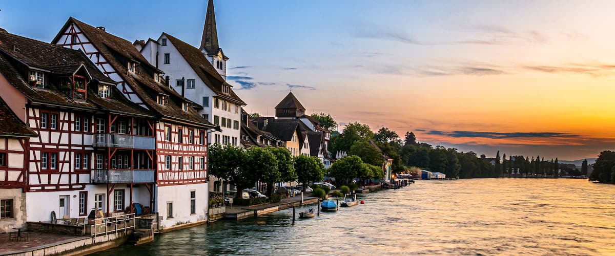A riverside town with traditional half-timbered houses, a church steeple, and a sunset over the water.